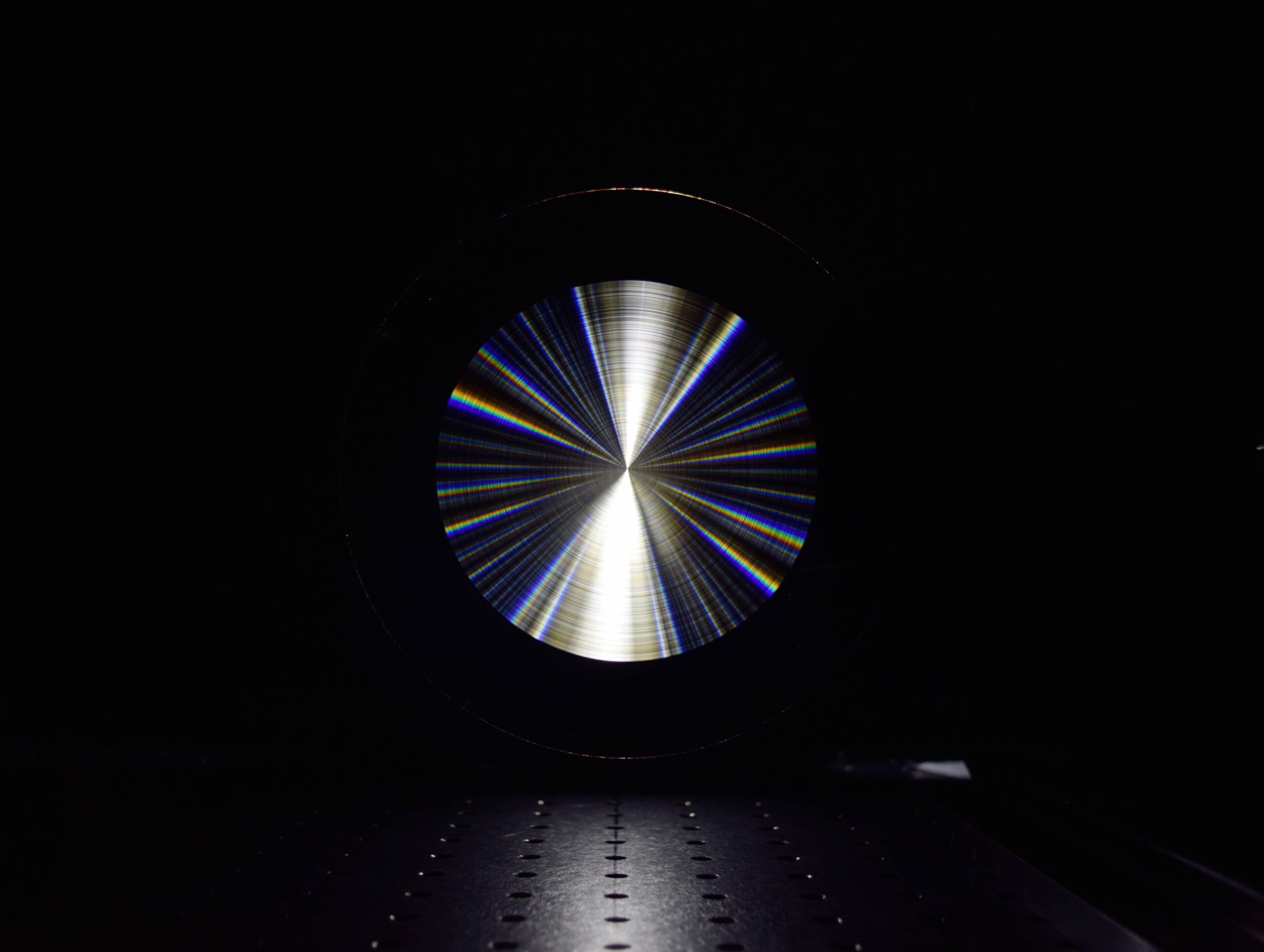 A flat disc lens in the dark reflecting multiple wavelengths of light.